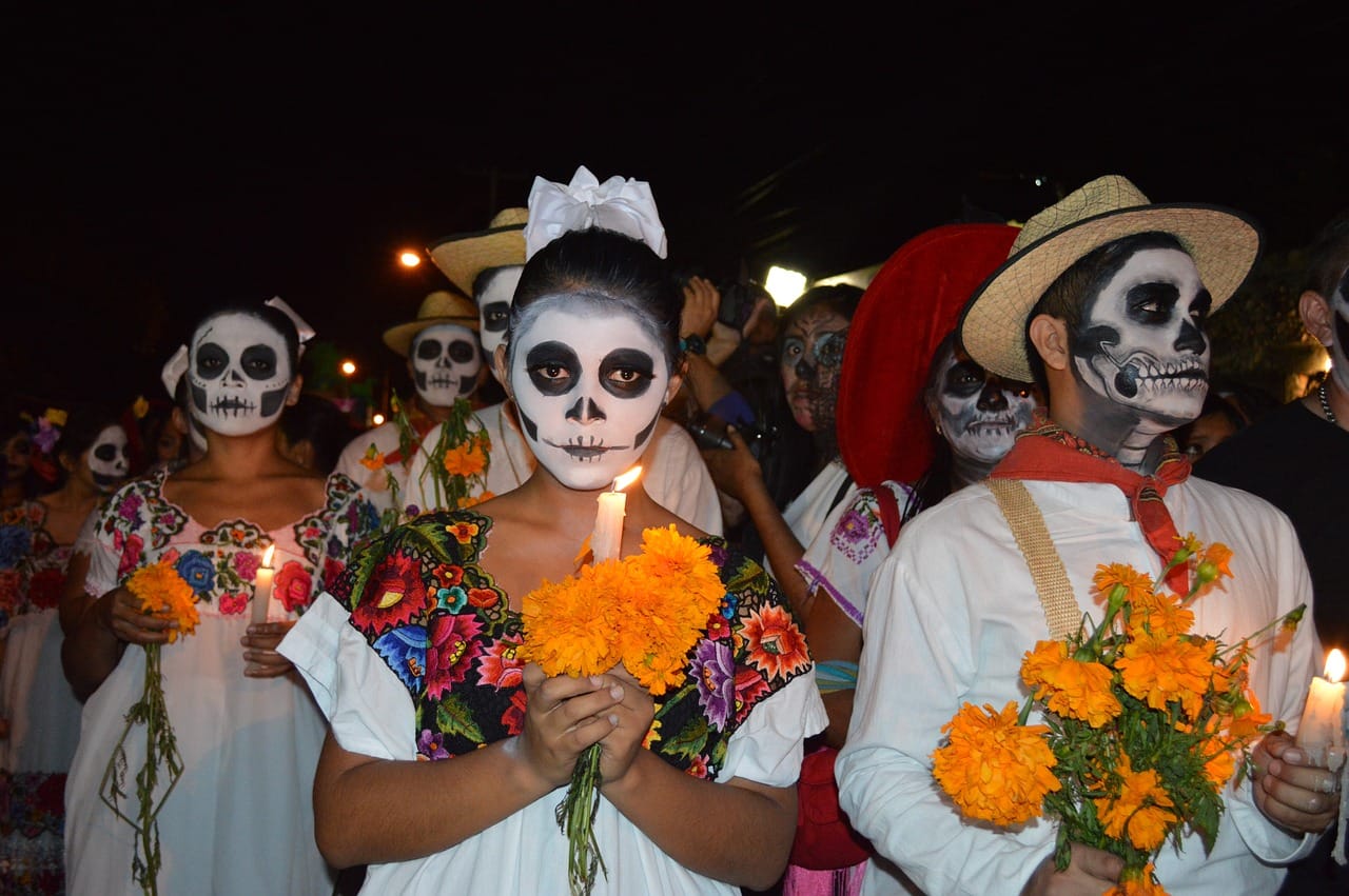 People have their faces painted like skulls. They're wearing white outfits and some are carrying orange flowers and lit candles.