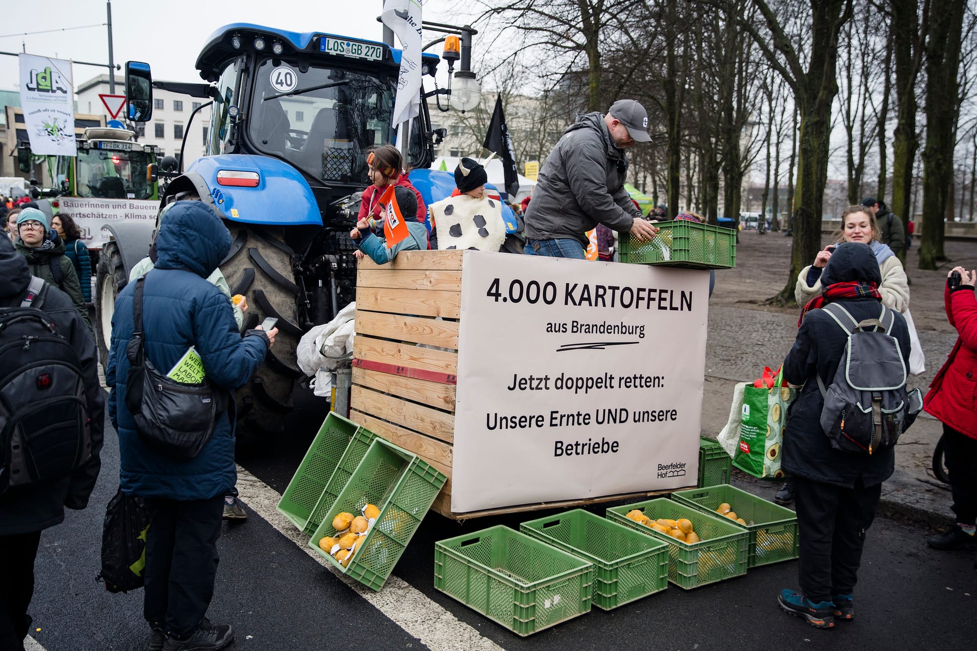A farmer has built a station next to a tractor to give out potatoes at a demonstration. People are around taking pictures, on their phone, or have potatoes in hand.