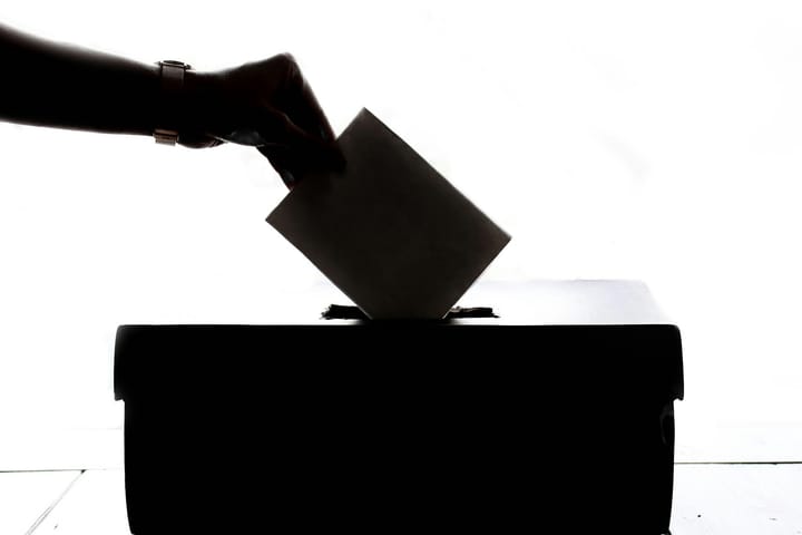 A person is dropping a ballot into a ballot box, all of which is black/shadowed. The background is white.